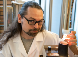 Dave is a white man with long dark hair and a beard. He's wearing glasses and a white lab coat which he works with some scientific equipment.
